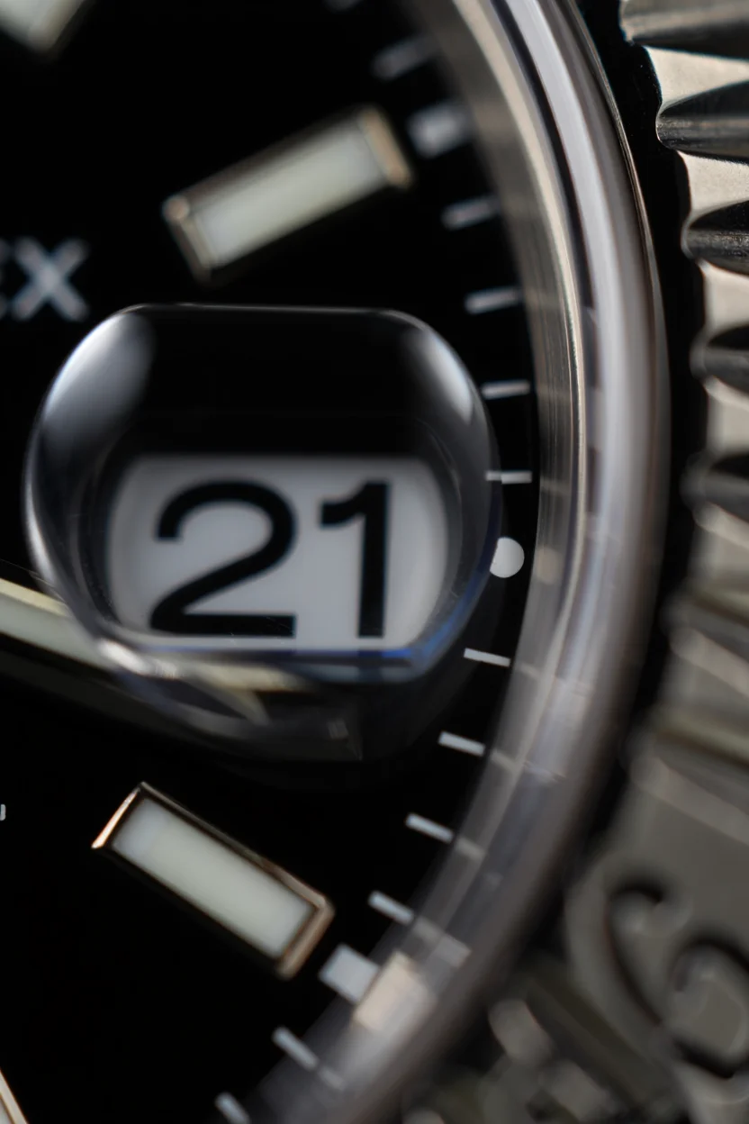 Ultra macro close up of the date window on a Rolex watch, showcasing the circular magnification lens over dark numbers on a black dial, with the characteristic cyclops lens creating an enlarged view of the date display with the number "21". The metallic bezel and surrounding watch face elements are visible at the edges of the frame, rendered in sharp detail against a blurred background. Ultra macro photography with shallow depth of field and sharp focus on the main subject.
