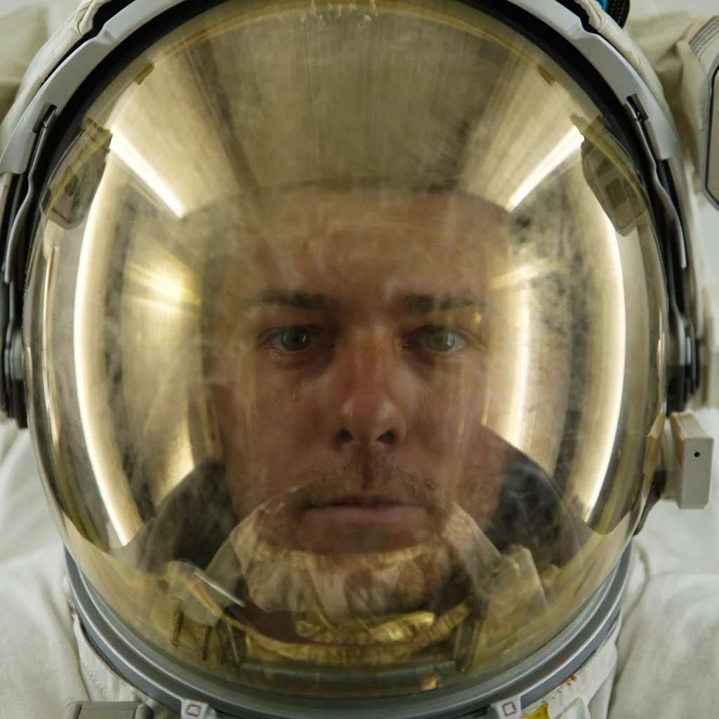 A close-up photograph of an astronaut's helmet with a reflective mirrored surface shows the astronaut's eyes barely visible through the highly mirror reflective surface. The helmet is a standard NASA-style space helmet with a curved gold-tinted visor that reflects light. The visor spans the entire width of the helmet, with the astronaut's eyes appearing as two small dark points through the reflective surface. Close-up macro photography with high-contrast lighting on reflective metallic surfaces.