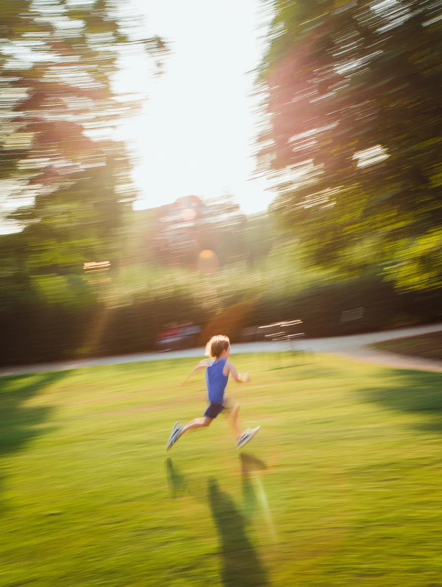 A still from a Super 8 home movie, showing a child running through a park on a brightly lit summer afternoon. The image exhibits the characteristic traits of Super 8 film, including noticeable film grain, flickering light, and slightly faded colors.  The footage is slightly overexposed with a warm color temperature, imbuing the scene with a nostalgic and dreamlike quality.