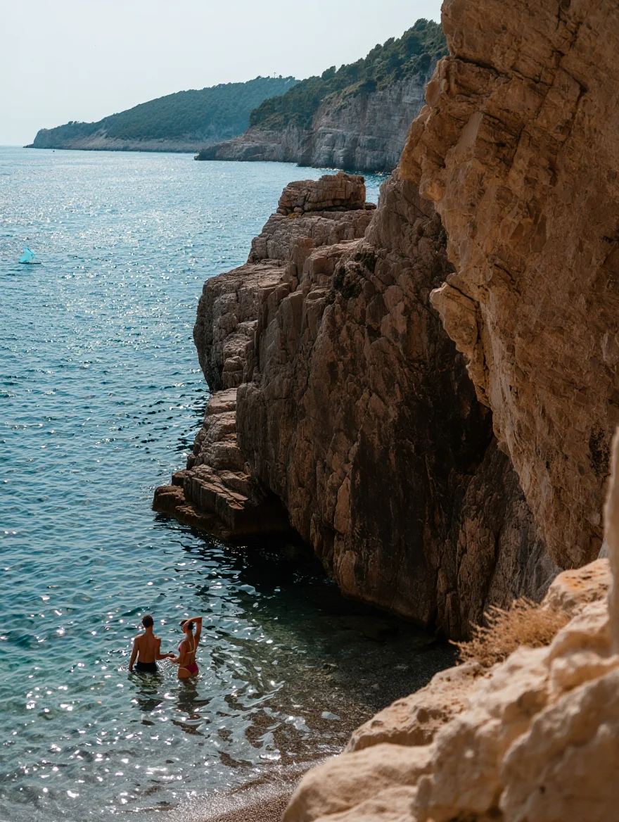 A scenic Mediterranean coastal landscape photographed in natural daylight from a high angle, featuring imposing limestone cliffs that rise dramatically from the crystal-clear blue-green water. Two swimmers wade in the sparkling sea near the base of the rugged cliff face. The background reveals a curving coastline dotted with vegetation-covered hillsides, while the cool color palette emphasizes the natural beauty of the scene. The composition is right-heavy, dominated by the textured rock formations, while the wide shot captures the grand scale of the coastal environment.