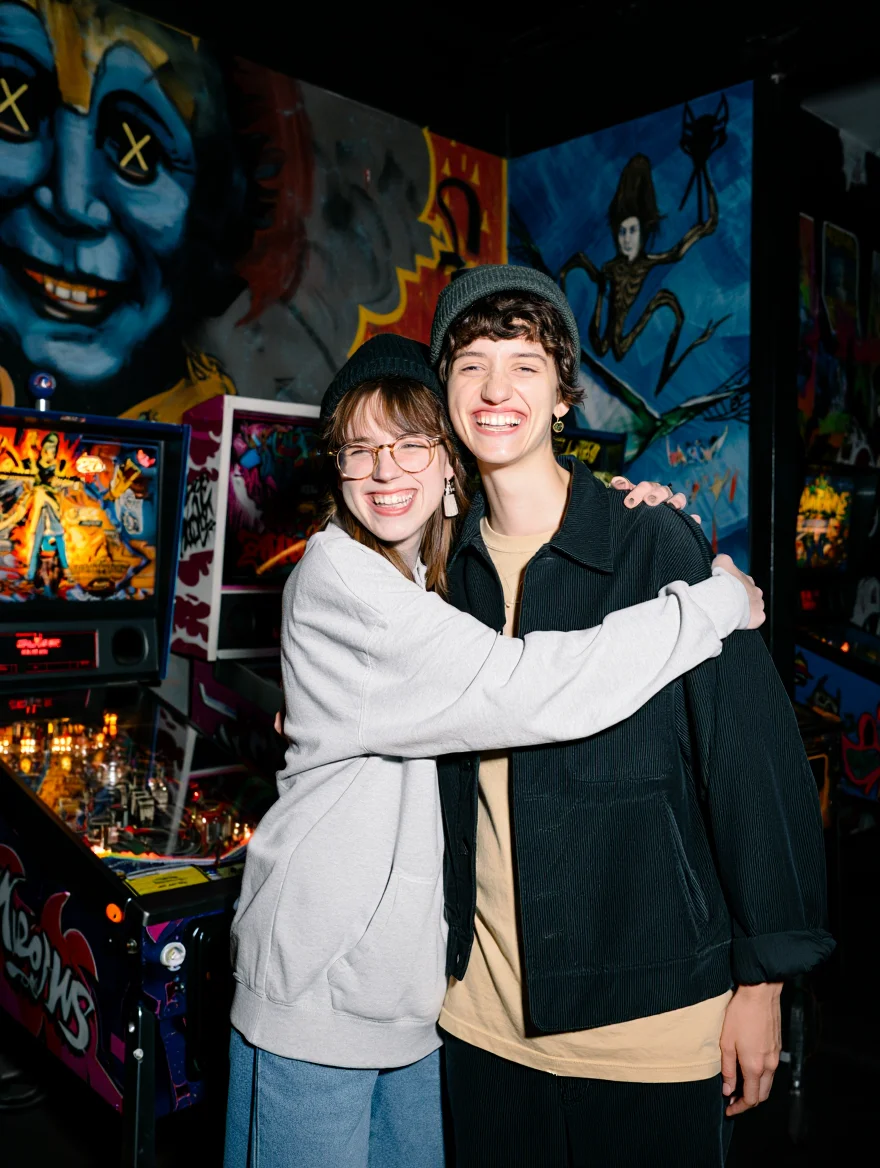 Two women embrace each other in a dimly lit arcade bar. One woman wears glasses and a light sweater with a beanie, while the other woman wears a dark corduroy jacket over a beige top and a beanie. Both women display warm smiles. Behind the women, a horror-themed pinball machine illuminates the space with its glowing display. The walls feature colorful graffiti artwork, including a large blue face with X-shaped eyes, alongside various abstract paintings. The scene is bathed in the bright flash that illuminates the dark space, highlighting the graffiti-covered walls and creating sharp contrasts. Indoor film photography with bright flash lighting.