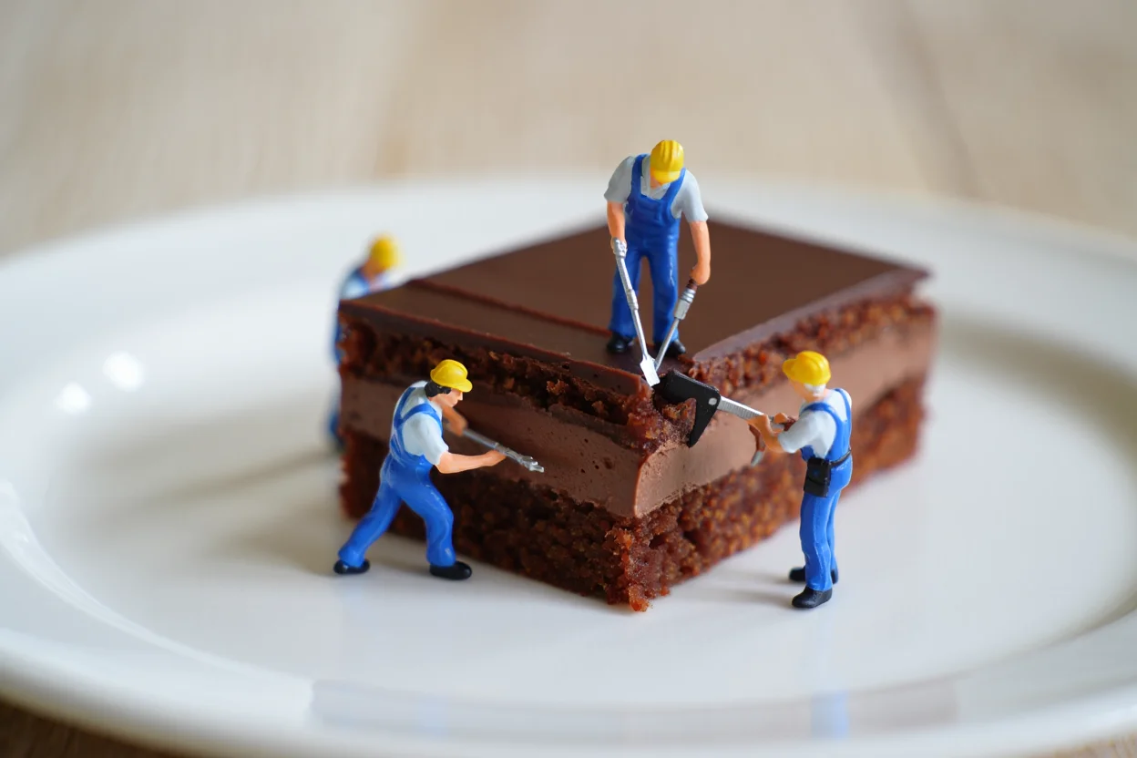 Several miniature worker figurines, approximately one inch tall, are positioned around a chocolate gateau on a white ceramic plate. The workers wear blue construction uniforms and yellow hard hats, appearing to repair or construct the chocolate cake using tiny tools. The gateau features multiple layers of dark chocolate frosting with a smooth, glossy finish. The scene is set against a soft, blurred background in neutral tones, emphasizing the detailed miniature scene in the foreground. Professional product photography with shallow depth of field and studio lighting against a soft-focus background.
