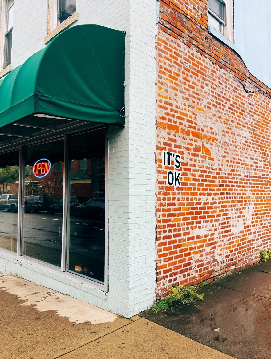 An urban street scene featuring the corner of a weathered brick building. The front-facing wall is painted white and features a forest green canvas awning extending over a glass storefront window. A red neon 'OPEN' sign glows in the window. The side wall reveals exposed red brick with simple text art reading 'IT'S OK' in black capital letters. The concrete sidewalk is wet from recent rain, creating reflections of the building and signage. Natural daylight illuminates the scene, casting soft shadows under the awning. Urban street photography with natural lighting and sharp focus on architectural details against a wet concrete surface.