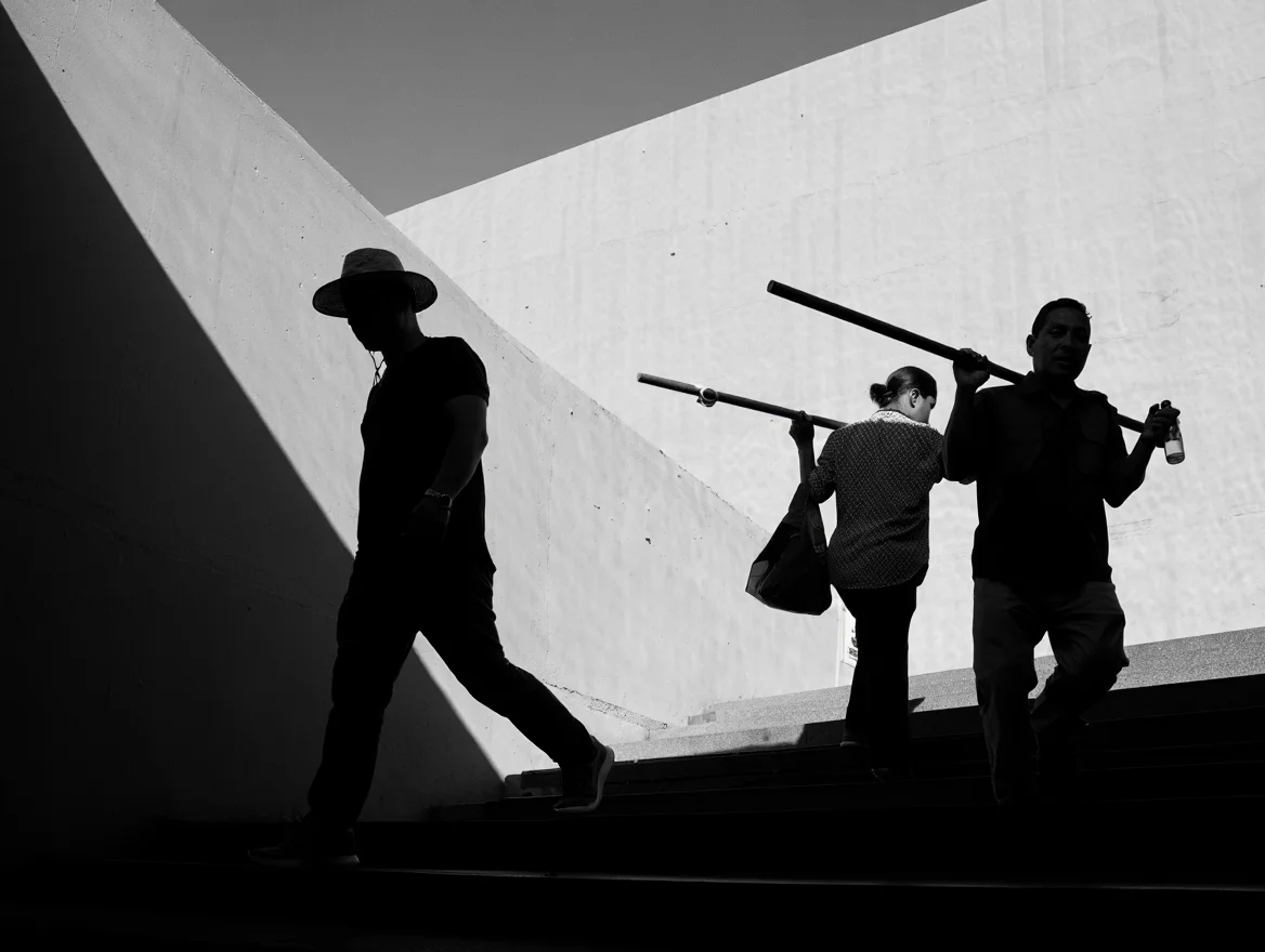 A striking black and white street photograph capturing three people ascending a diagonal architectural ramp. The composition emphasizes strong geometric lines and dramatic contrast between light and shadow. The scene features a lead figure silhouetted in dark clothing and a wide-brimmed hat, followed by two people carrying poles across their shoulders. Natural side lighting creates bold shadows across the white architectural surfaces, while the high angle perspective emphasizes the diagonal movement through the frame. The deep depth of field ensures all elements are in sharp focus, from the architectural details to the human subjects.