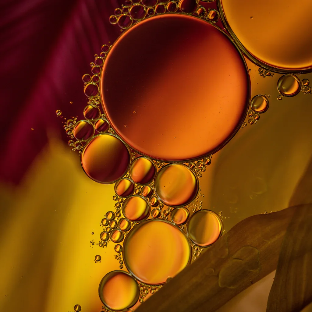 Extreme macro photograph capturing the mesmerizing interaction between oil and water, showcasing cellular-like structures formed by surface tension. The image features multiple translucent oil bubbles of varying sizes floating on water, creating distinct boundaries where they meet. The bubbles display a warm color palette dominated by amber, gold, and burgundy tones, enhanced by artificial side lighting that accentuates their translucent quality. The composition is balanced with both large and small bubbles distributed across the frame, while the shallow depth of field creates a dreamy, abstract quality. In the background, blurred plant material and colored surfaces in burgundy and golden yellow add depth and contrast to the composition. The extreme close-up perspective and macro lens capture the intricate details of this natural phenomenon, revealing the beautiful patterns and textures created by the physical properties of these immiscible liquids.