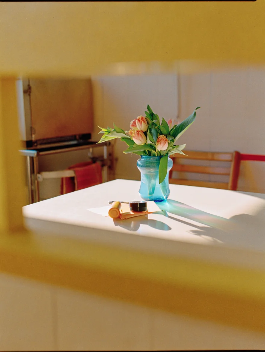 A still life on a kitchen table in the afternoon sun, shot on 1990s consumer-grade film stock. The image features visible film grain, slightly faded colors with a warm tint, and minor light leaks around the edges. The photograph exhibits soft focus and subtle vignetting, typical of low-cost camera lenses from that period.