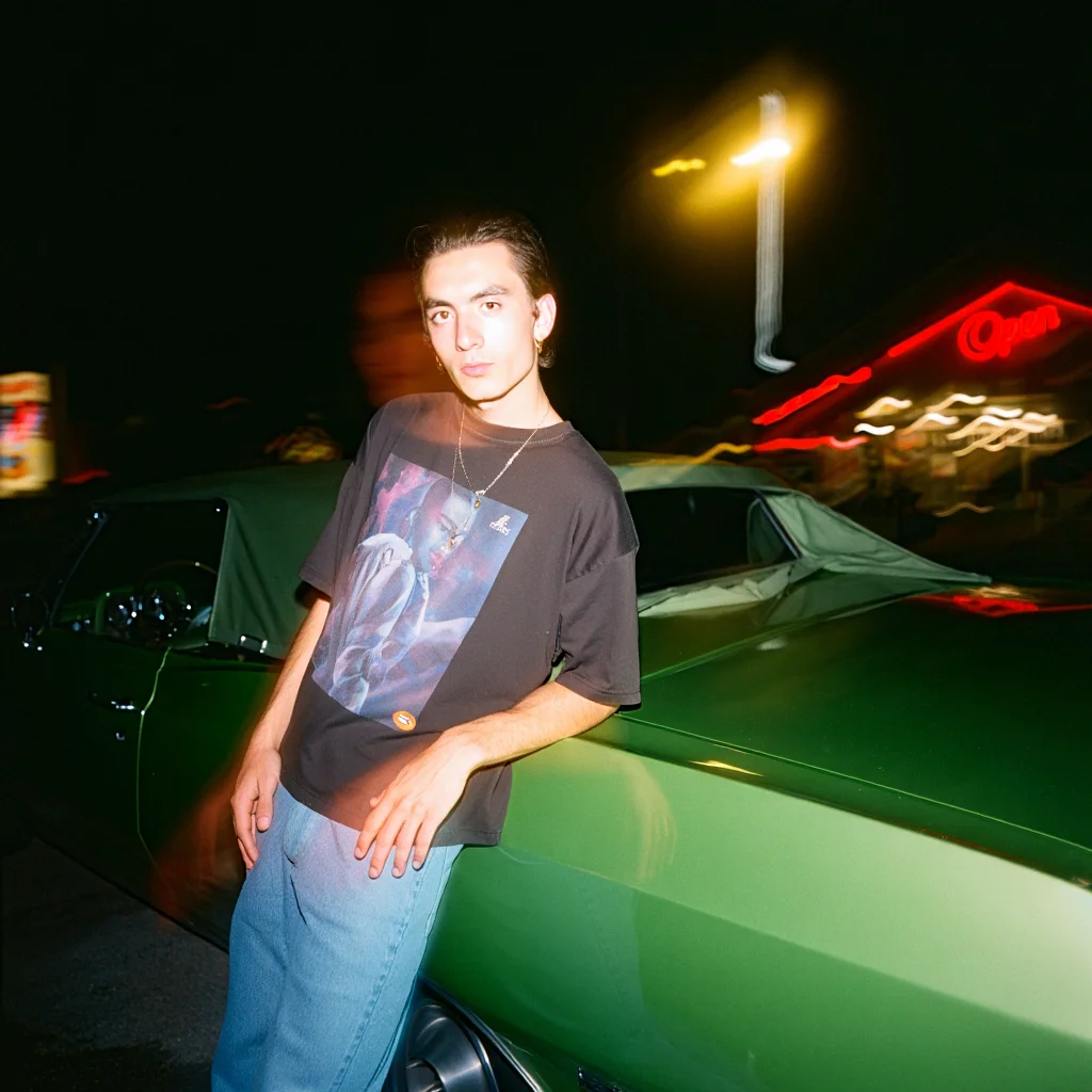 A blurry Y2K photograph capturing a man with frosted tips wearing a baggy graphic tee and jeans. He leans against a green vintage car on a nighttime street, illuminated by the warm glow of a streetlight. The background is blurred, showing the hazy lights of a diner and a neon "Open" sign. The image has a grainy texture, resembling a scanned photo taken with a disposable camera.