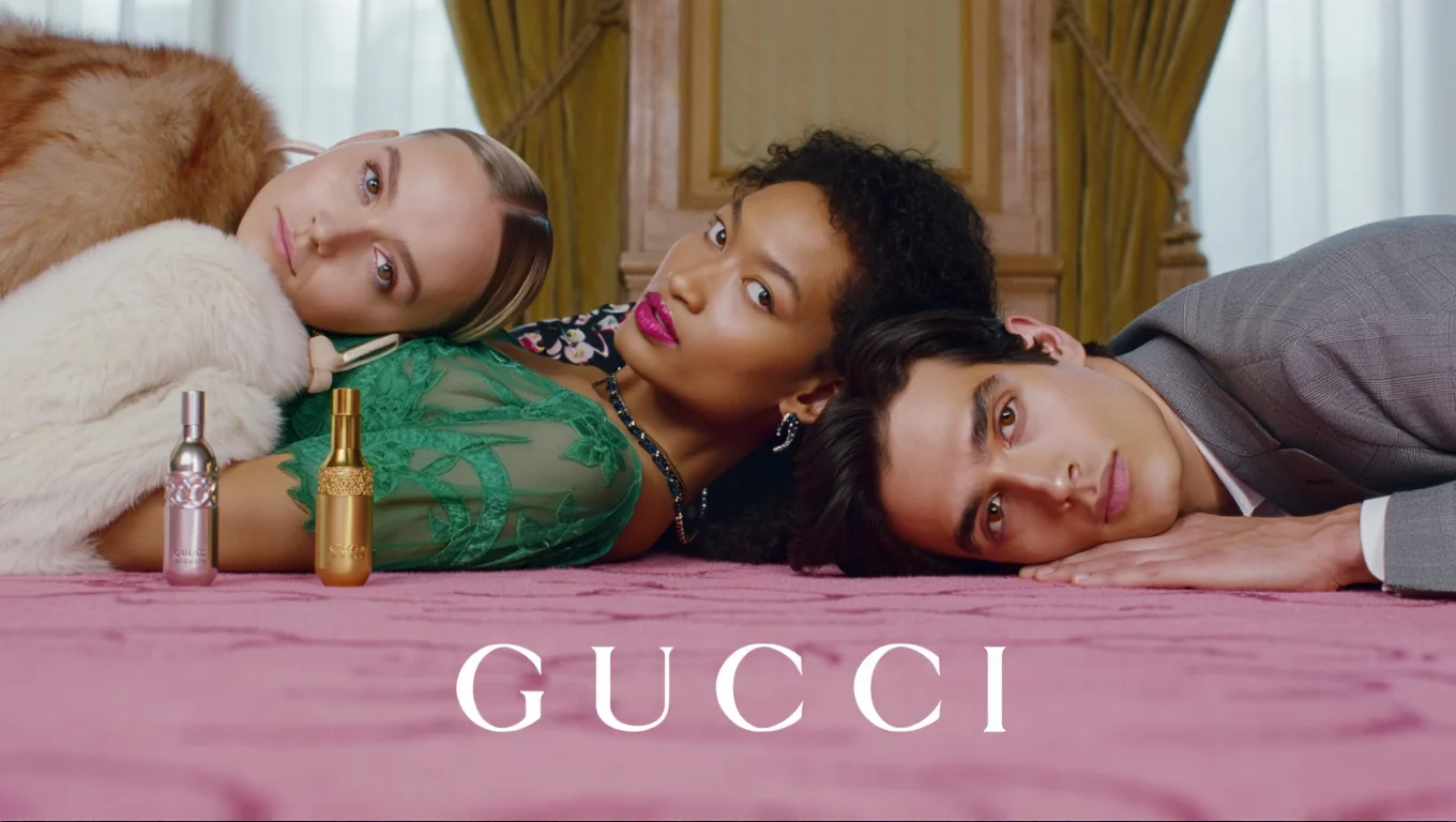 A luxury fashion campaign photograph for Gucci fragrances, shot in an elegant interior setting with pink carpeting and gold curtains. Three models are artistically arranged on the floor with their faces clearly visible, including one wearing a green lace dress with a white fur coat, another in a black floral print shirt, and a third in a grey suit. Two Gucci perfume bottles, one gold and one silver, are prominently featured. The scene is captured from a three-quarter angle with professional studio lighting creating an even, luxurious atmosphere. The white "GUCCI" logo appears at the bottom of the image in serif capital letters. Professional fashion photography with clear facial features and studio lighting.