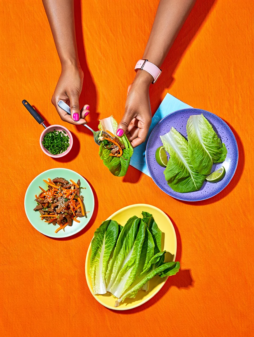 An overhead view of a vibrant Asian-inspired lettuce wrap feast arranged on a bright orange textured surface. The composition features multiple colorful plates: a mint green plate with stir-fried meat and shredded carrots sprinkled with sesame seeds in the lower left, a yellow oval plate with fresh romaine lettuce leaves in the bottom center, and a purple-blue speckled plate containing assembled lettuce wraps garnished with lime wedges on the right. A small pink bowl with fresh green herbs sits in the upper left, accompanied by a serving utensil. A pair of medium-toned hands enter the frame from above – the left hand holding a spoon with bright pink nail polish visible, and the right hand displaying a freshly assembled lettuce wrap. The left wrist features a light pink watch. A light blue napkin peeks out from under the purple-blue plate, adding to the colorful arrangement. Overhead food photography with bright direct lighting and defined shadows on a solid colored background.