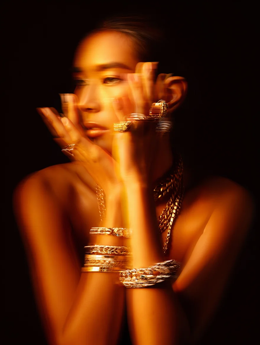 A high-fashion editorial photograph for Numéro magazine featuring a side profile of a model adorned with multiple pieces of luxurious gold jewelry. The image employs dramatic side lighting creating a warm, golden glow, with intentional motion blur effects that add a dreamy quality. The composition is enhanced by the interplay of light on the jewelry pieces, including bracelets, rings, and necklaces, all captured against a dark, moody background. The aesthetic is elevated by artistic lighting techniques that create striking reflections and shadows.