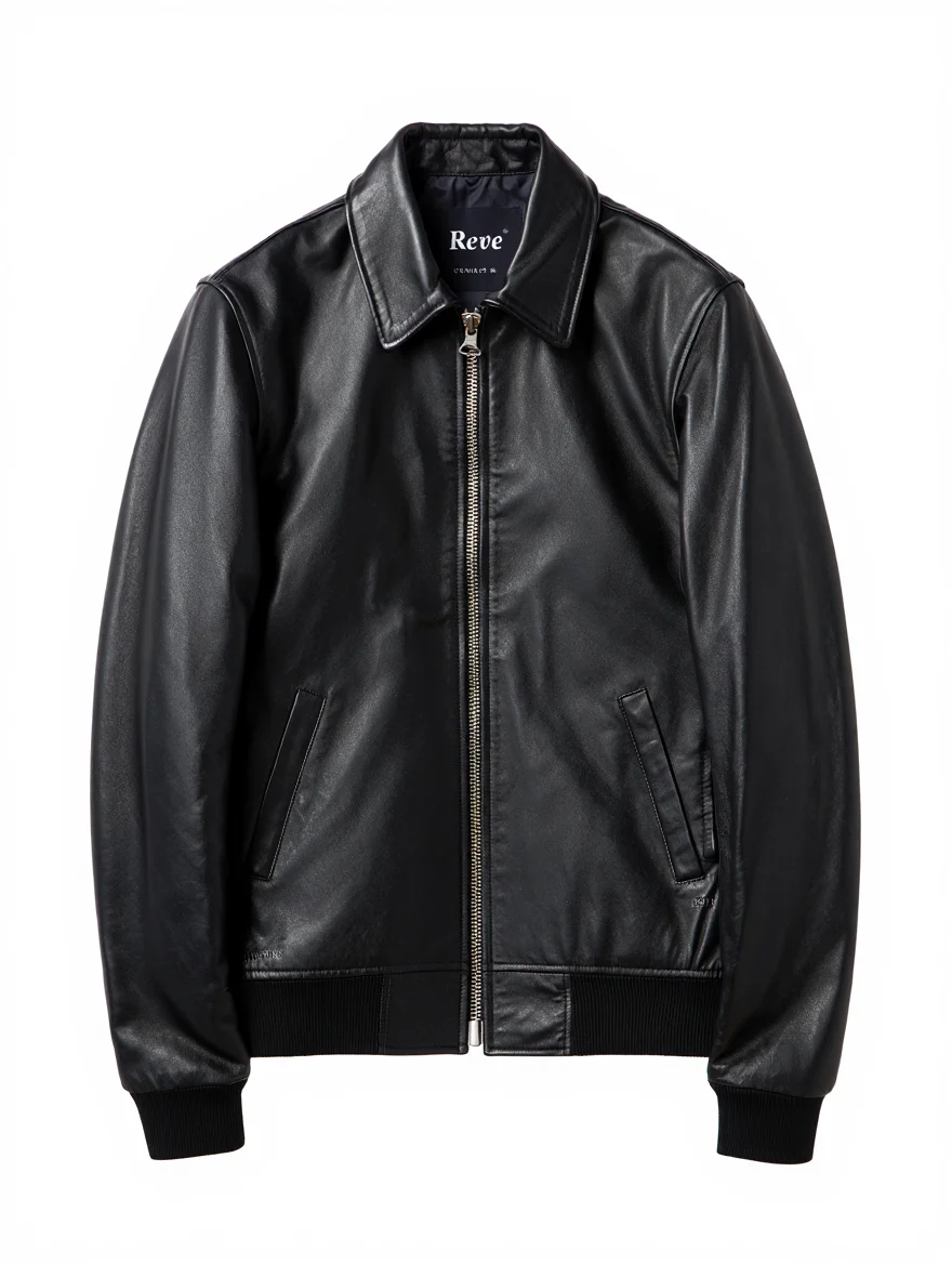 A product photograph captures a classic black leather bomber jacket centered against a pure white background. The jacket features a pointed collar, silver metal zipper front closure, two angled front pockets, and black ribbed knit cuffs and waistband. The leather material displays a distressed, vintage look with subtle sheen under studio lighting. The composition is perfectly centered with even, front-facing artificial lighting that highlights the jacket's premium construction and details. A tag inside the jacket displays the text "Reve" in a distinctive swash font.  Professional product photography with studio lighting setup and clean white background composition for commercial use.
