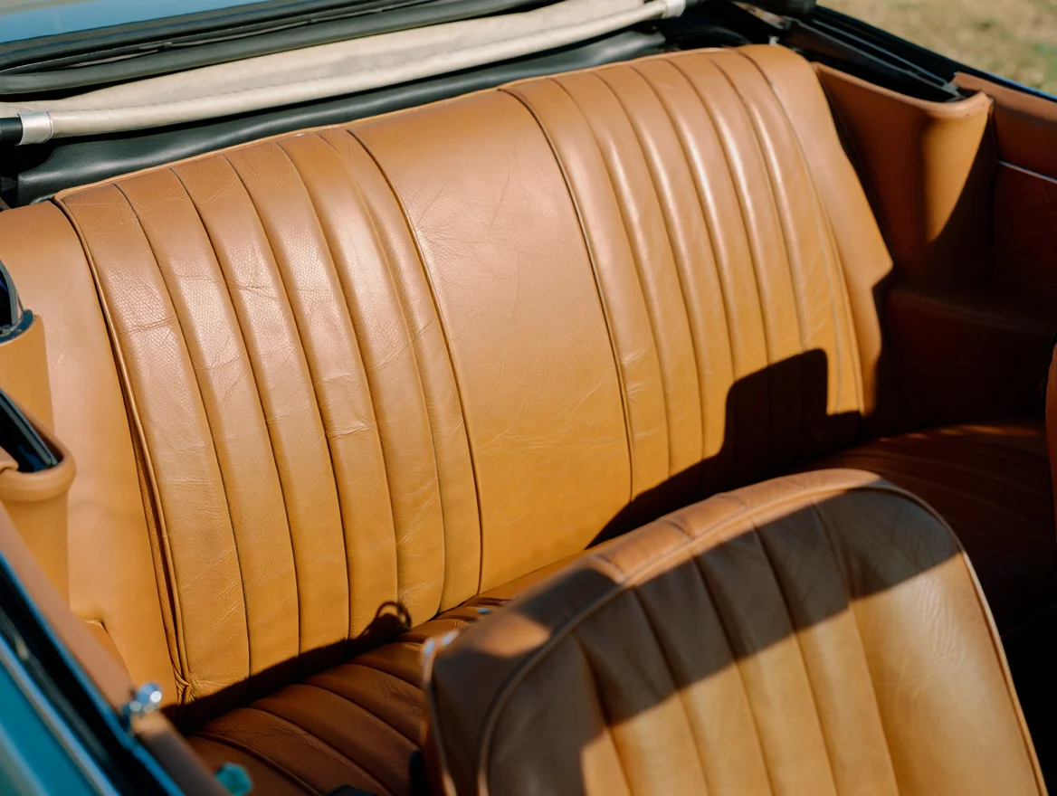 A vintage film photograph taken from inside a convertible car shows the tan leather backseat with visible stitching and weathered creases. The folded fabric roof is partially visible above, creating a natural frame around the composition. Warm sunlight illuminates the worn leather surfaces, highlighting the authentic patina and texture of the material. The backseat spans the full width of the frame, with subtle shadows cast by the interior components.  Vintage 35mm film photography with natural grain texture and soft ambient lighting.