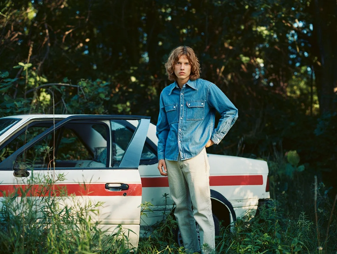 A casual snapshot shows a figure wearing a denim shirt and light-colored pants standing with one hand on hip next to a partially visible white car with red striping, the car appearing wrecked or abandoned among overgrown brush. Natural daylight filters through trees in the background, casting dappled shadows across the scene. Wild grass and foliage grow in the foreground around the car. The photograph exhibits characteristic 1990s film qualities including muted colors with blue-green color shifts in shadow areas, subtle film grain texture, and slightly soft focus throughout the frame.  Amateur film photography from the 1990s with natural lighting and characteristic film color rendering.