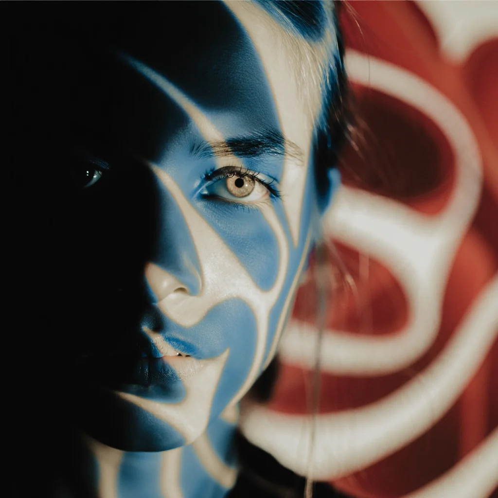 An ethereal blue and sepia toned portrait, centered on the subject's eye peering from the left side of the frame, partially obscured by shadow.  The composition uses a shallow depth of field, with the background a swirl of abstract shapes in deep red and white. The overall effect is one of mystery and intrigue, as if the subject is looking out from a dream.