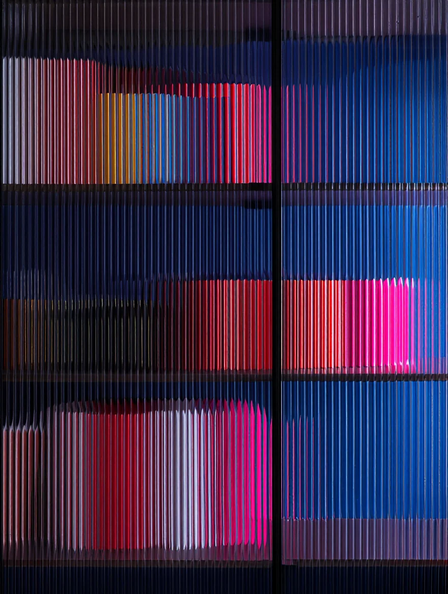 Thick ridged glass with pronounced texture distorts and blurs an array of objects behind it, creating an abstract pattern of vertical color blocks in deep blues, vibrant pinks, and rich reds. The glass surface fills the entire frame, with its wavy texture creating a ribbed effect that fragments and refracts the colors behind it into elongated stripes. Close-up macro photography with sharp focus on textured glass surface in controlled indoor lighting.