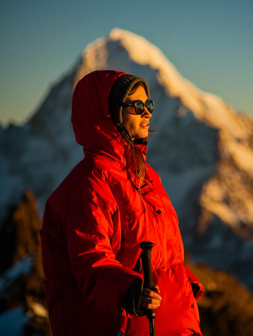 A lone female hiker pauses on a ridge line in the Andes, photographed in a medium shot with a snow-capped mountain peak creating a dramatic backdrop. Bathed in the golden light of the setting sun, she stands facing the camera, wearing a bright red parka and sunglasses, while holding a trekking pole.  The image exhibits a warm color palette with a cinematic feel.