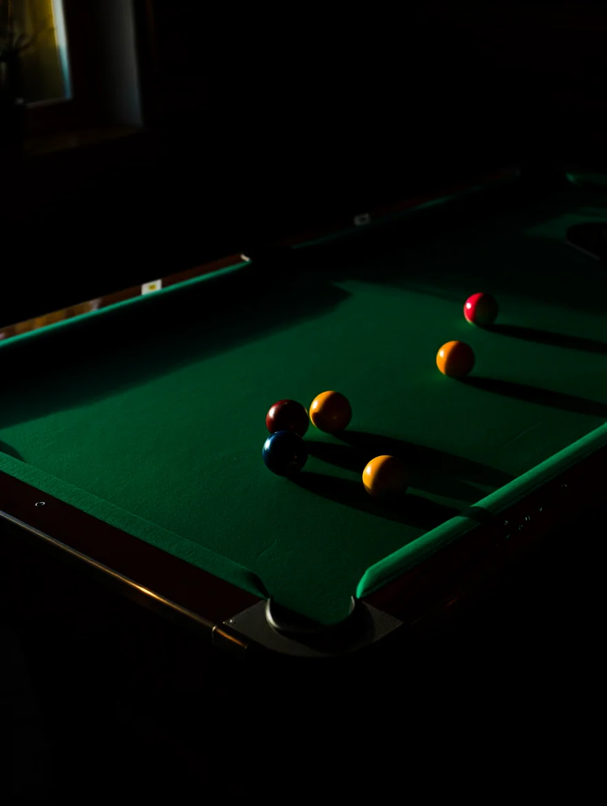 Several colorful billiard balls rest on a deep green felt pool table in a dimly lit bar setting. The glossy spherical balls cast distinct shadows on the table's surface, with some balls positioned near corner pockets while others cluster in the center. Dark wooden rails frame the table's perimeter, and the shadowy bar environment is visible in the background, with ambient lighting creating stark highlights on the balls' surfaces. The scene's dramatic lighting emphasizes the contrast between light and shadow across the curved surfaces of the billiard balls and the rich texture of the felt. High contrast film-inspired photography shot on Kodak Portra 400, with dramatic chiaroscuro lighting and deep shadows in a dark interior setting.
