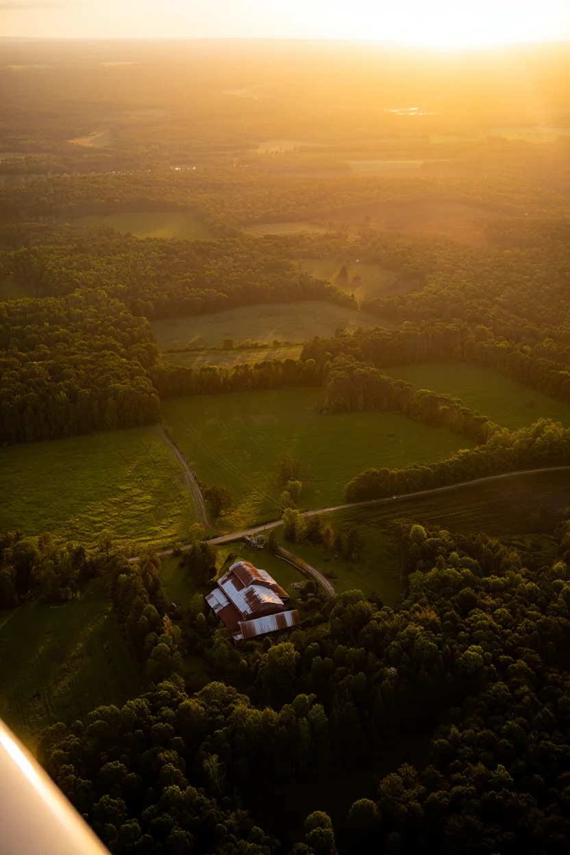 A vintage sepia-toned photograph, taken from a low-flying biplane, showcasing a sprawling patchwork of farmland and forest. A rustic barn, painted a faded red, stands out against the lush greenery, while dirt roads meander through the landscape, illuminated by the warm, hazy light of the setting sun. The image evokes a sense of nostalgia and the timeless beauty of rural life.