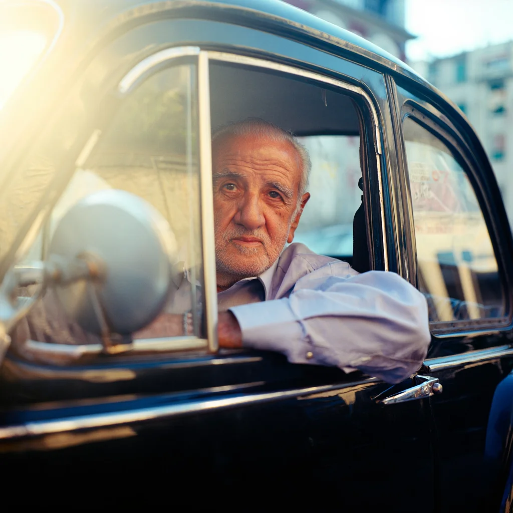 A portrait of an elderly Italian man in a classic black car parked on a sun-drenched street in Naples. The man's face is bathed in the warm, golden light of the late afternoon sun, his gaze distant as he rests his hands on the steering wheel. The scene evokes a sense of nostalgia and faded grandeur, captured on 35mm film with a slightly faded color palette and subtle light leaks.